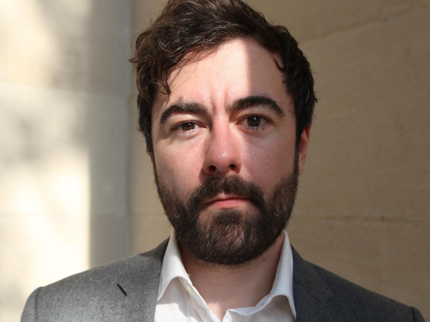White man with dark hair posing with a grey blazer and white shirt