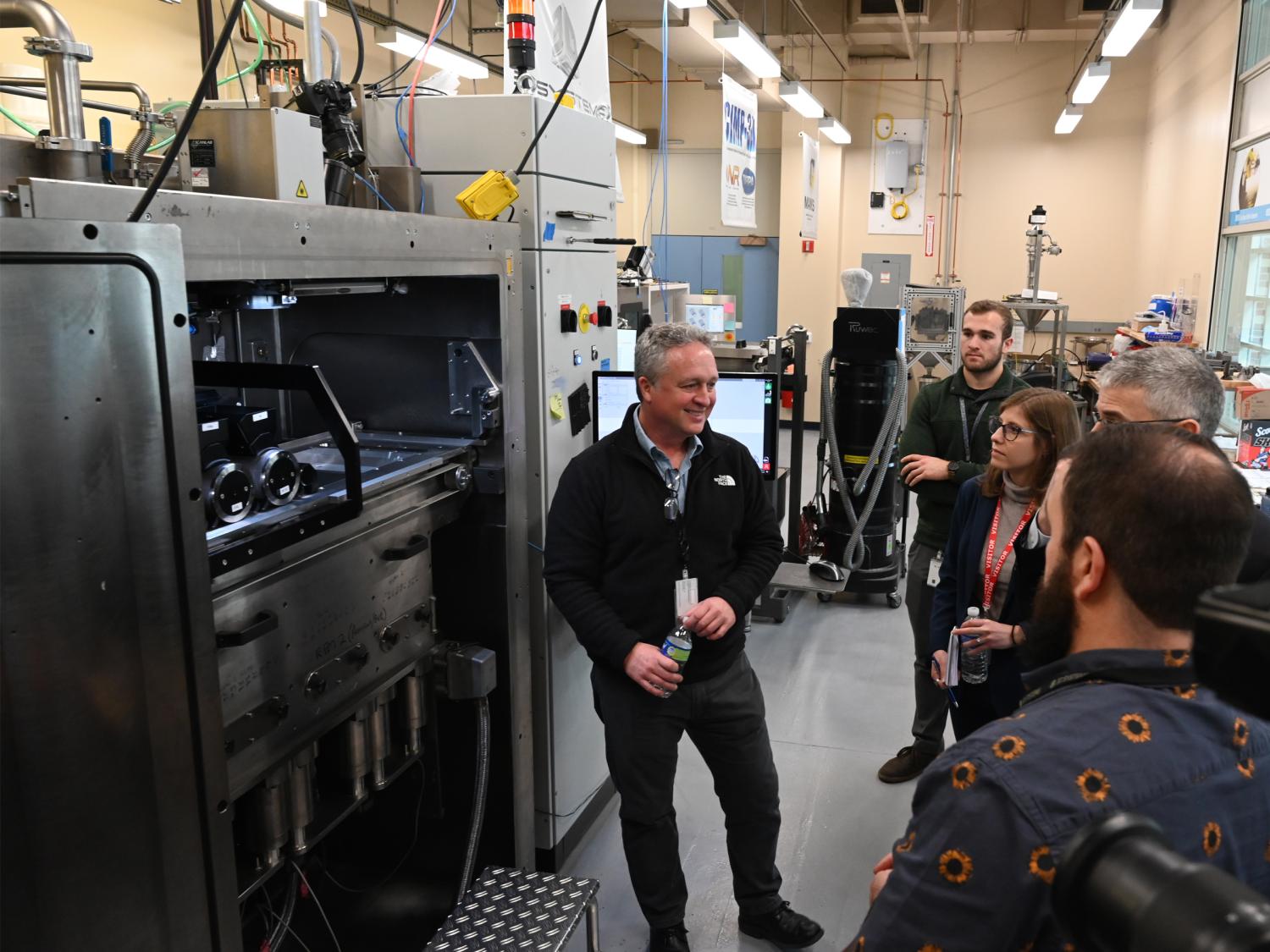 A small standing group of people listens to a presenter in front of lab equipment. 