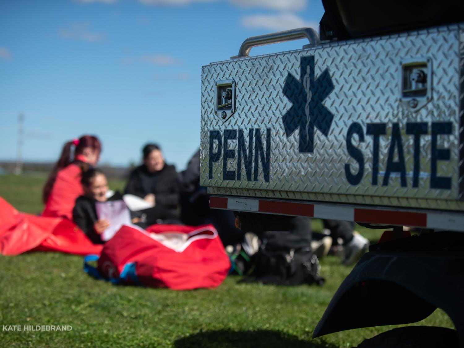 A Penn State emergency vehicle is parked on a grassy field with students sitting on the ground in the background.