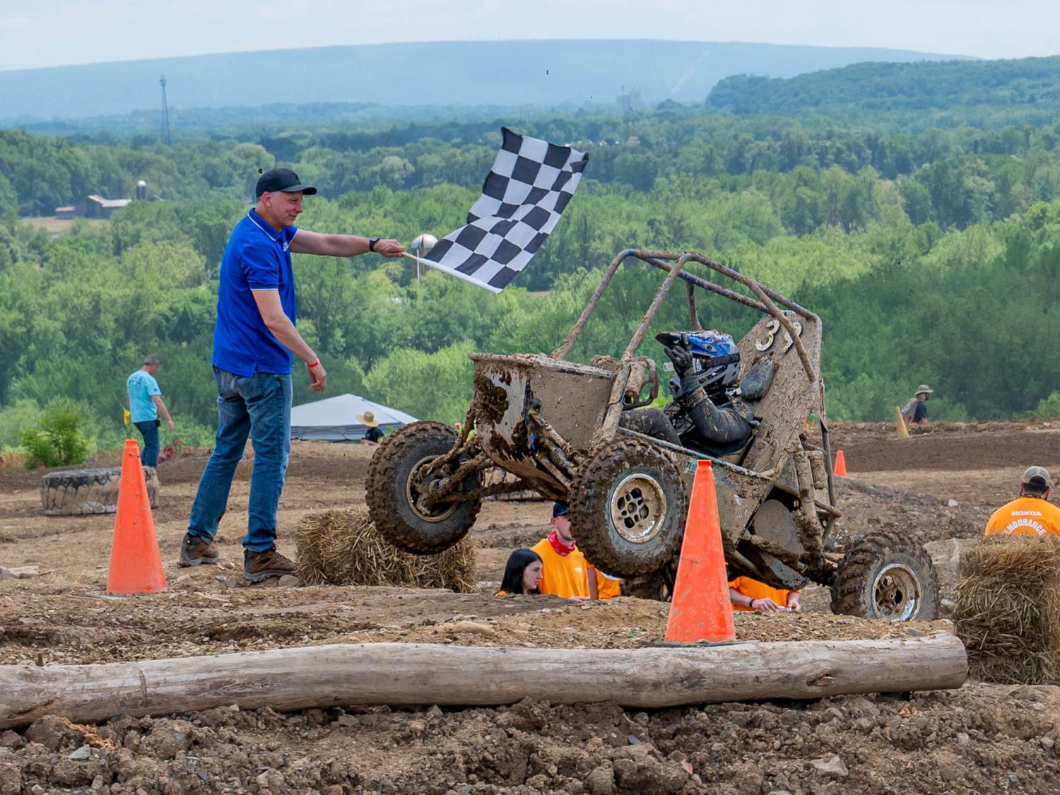 A man waves a checkered flag with an all terrain vehicle behind him