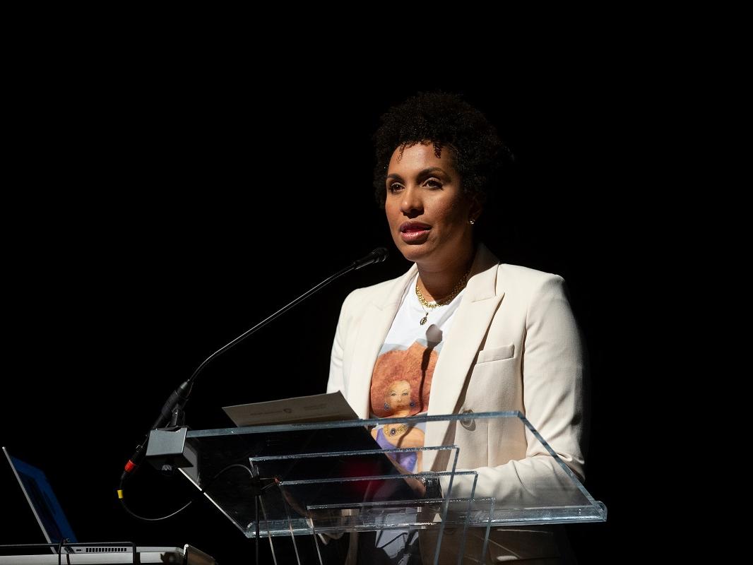 A woman stands at a glass lectern with a microphone that extends toward her. A piece of paper is visible on the podium. An open laptop sits on the table to her left.