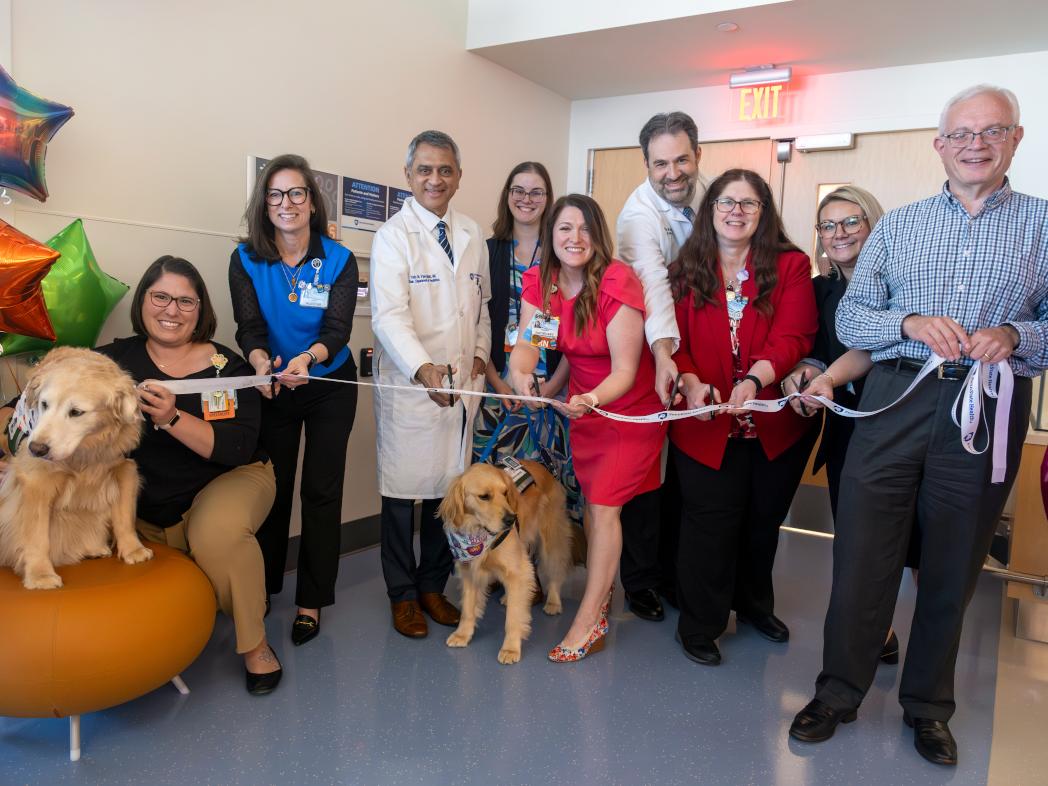 A group of people, and two dogs, smile for a photo before cutting a ribbon to open a new hospital unit