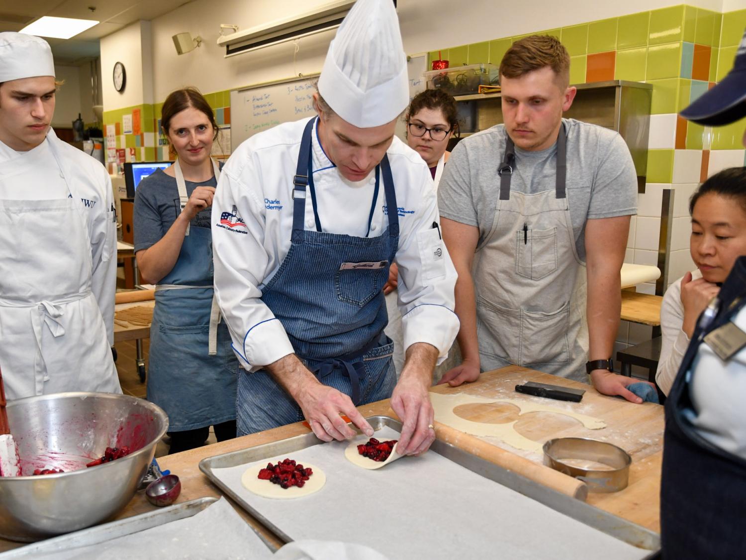 A man in chef's attire works at a table with students around him paying attention