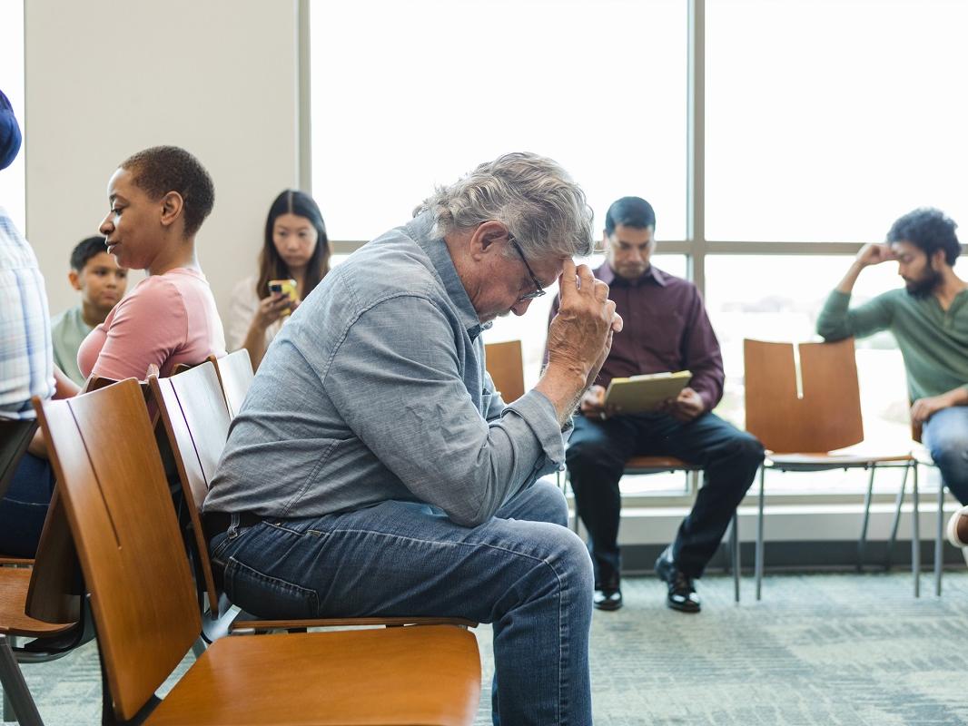 An older adult man sits anxiously in a hospital waiting room with his hands against his forehead.