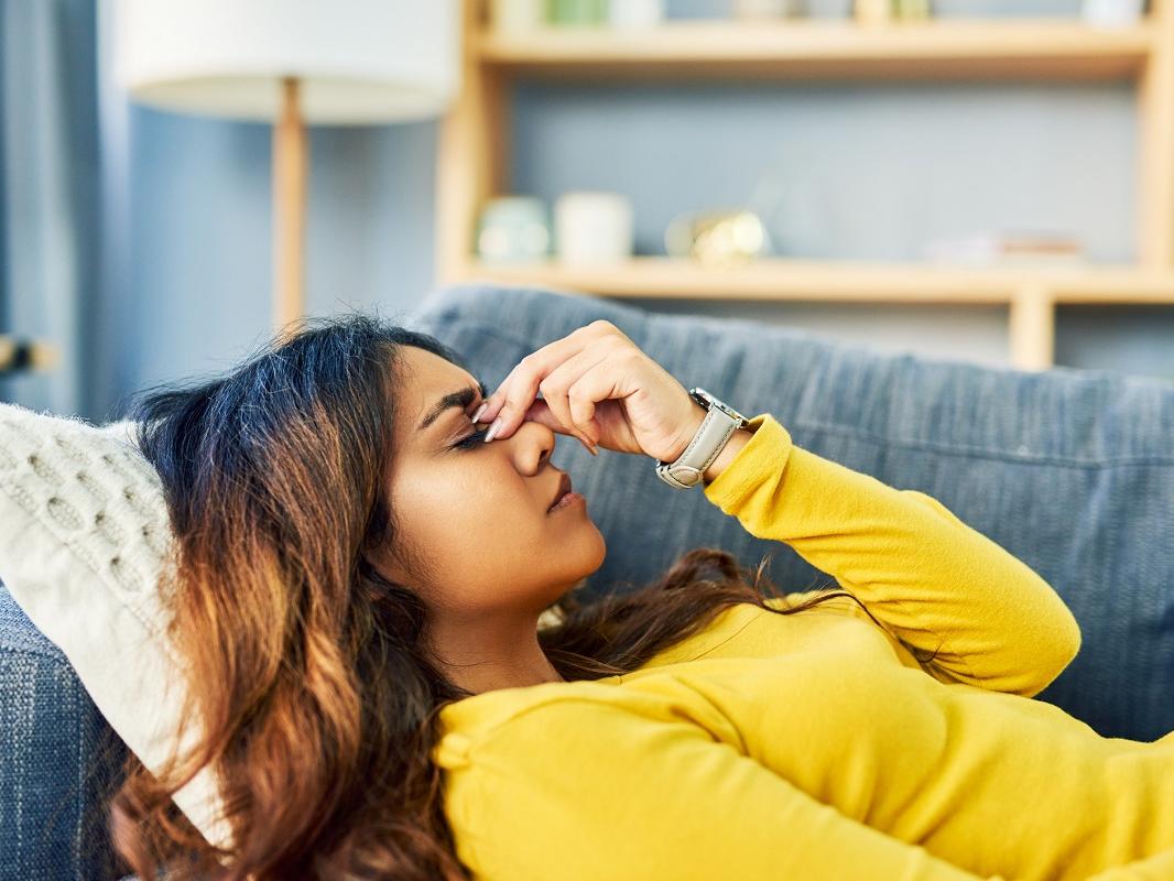 A woman lays down as she experiences a headache. She is lightly holding the bridge of her nose with her left hand and has her eyes closed.