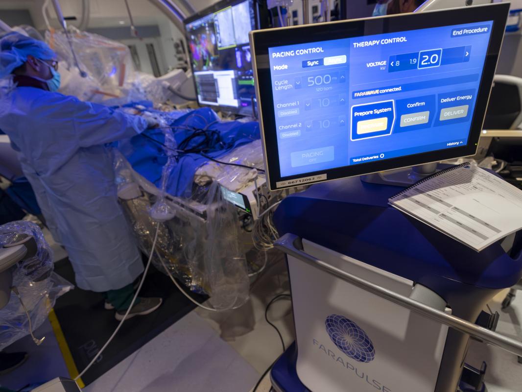 A physician wearing a cap, mask and gown performs a procedure on a patient, referencing a nearby monitor. Another monitor is located closer to the camera.