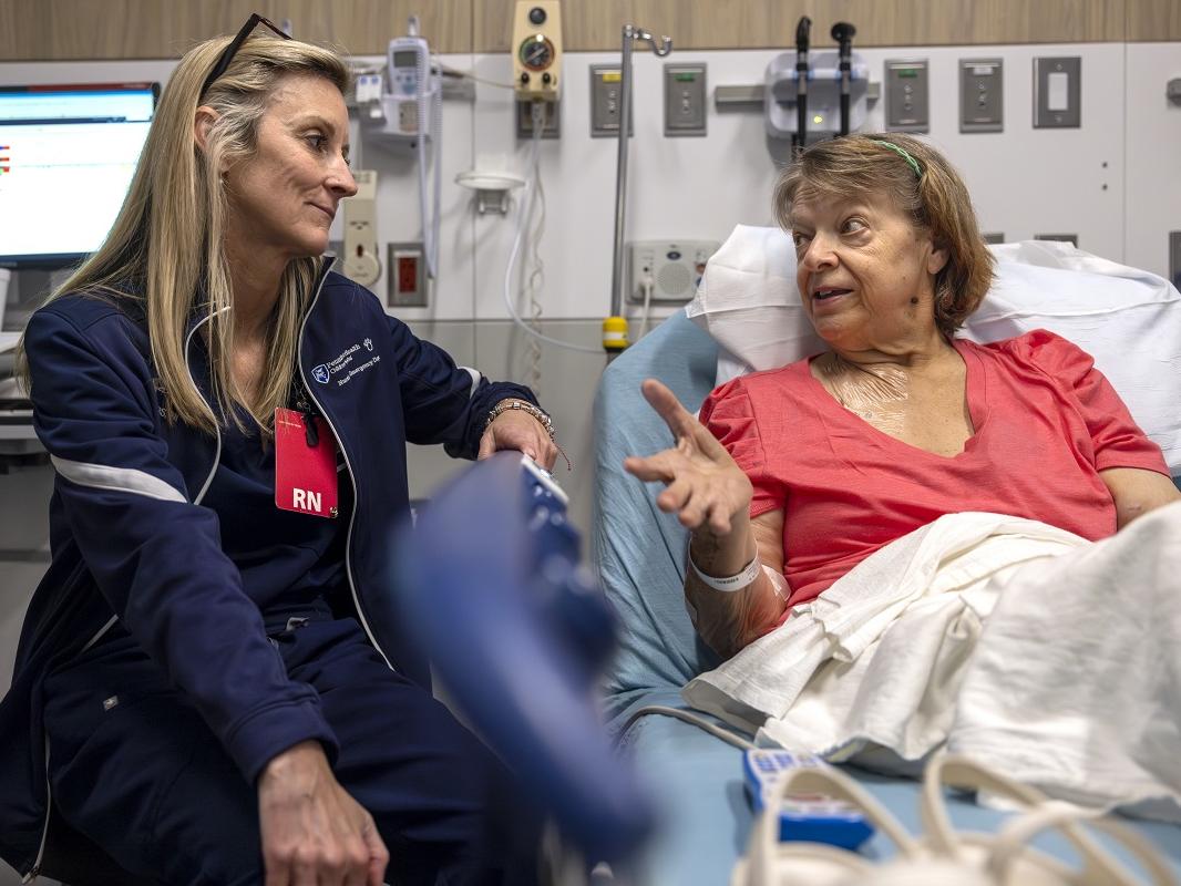 A female registered nurse sits by the bedside of a female patient in the Emergency Department as the patient talks to her. A computer monitor, medical equipment and monitors are behind the two women.