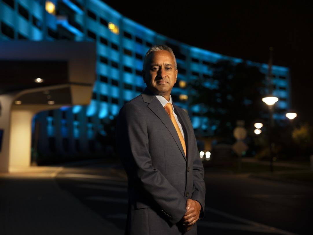 A man poses for a portrait in front of a building. The building is illuminated in blue light.