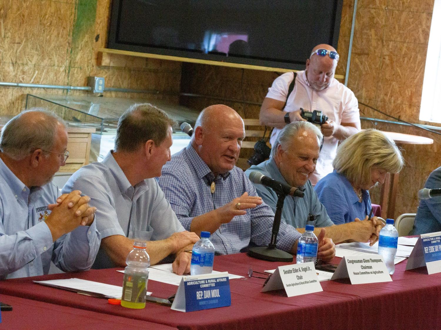 Five people sit at a table with a microphone. A sixth person is visible in the background. 