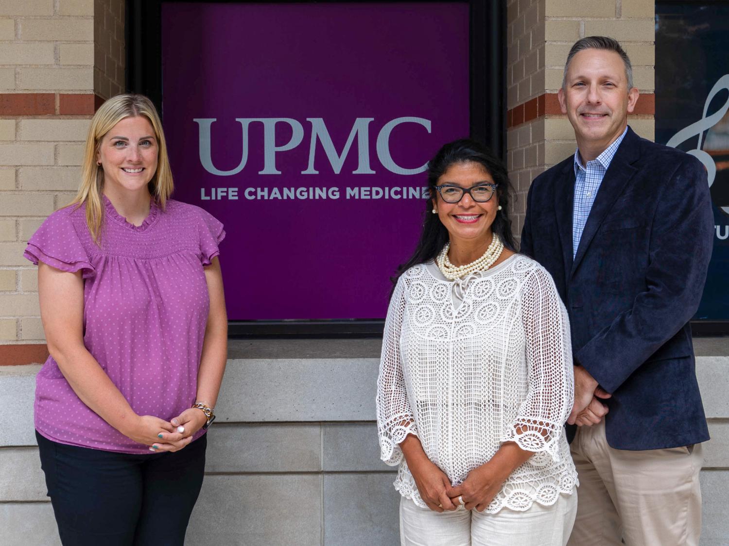 Three people pose for a photo in front of a sign that says UPMC