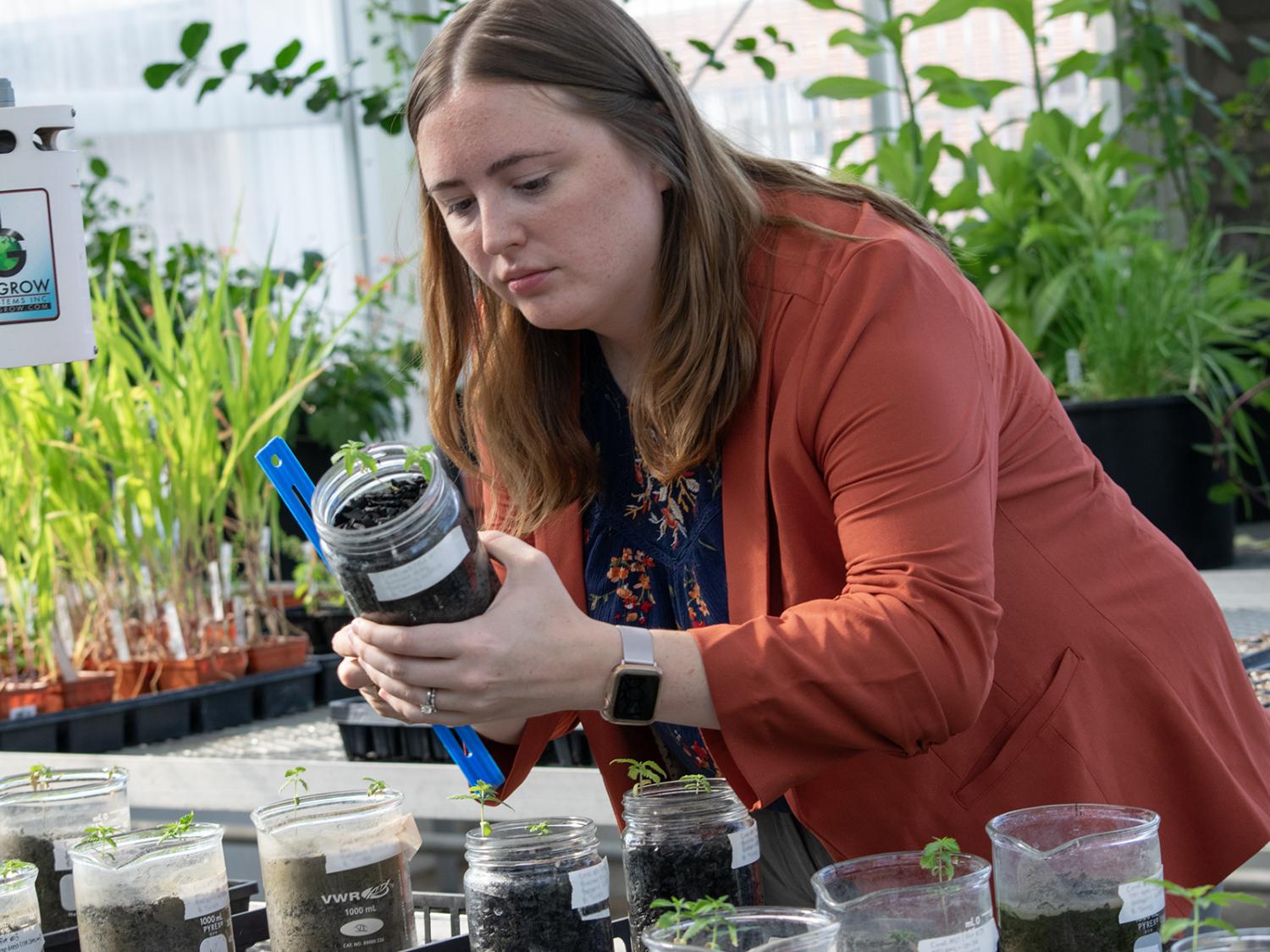 A woman holds a jar of soil and a ruler, taking a measurement, with plants behind her