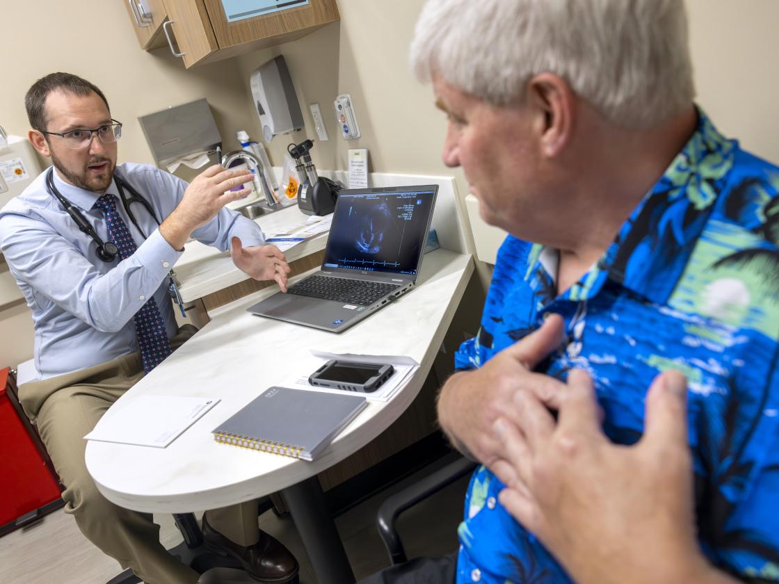 Tyler Thomas, left, gestures as he talks to patient John Jones, right. Thomas is wearing a button-down shirt, tie, glasses and has a stethoscope around his neck. Jones is wearing a Hawaiian shirt and has hands over his heart.