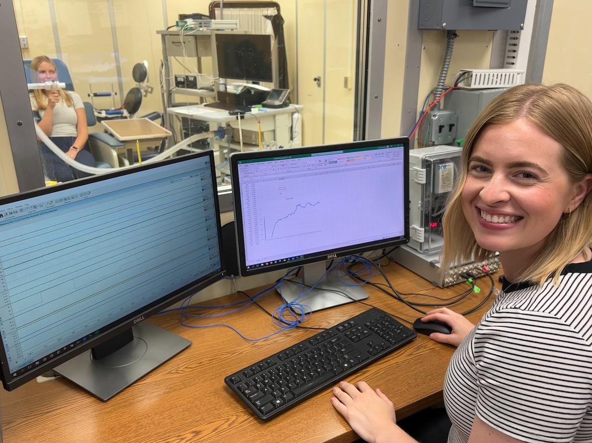 Olivia Leach smiles behind a bank of computer screens while Rachel Cottle sits behind a glass window