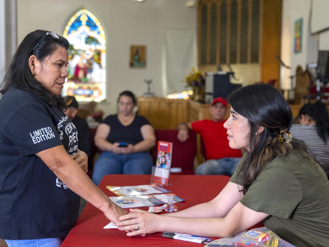 Two women hold hands as they speak to one another. One woman sits at a table and the other stands. They’re in a church, a stained glass window is behind them and people in a pew look on.