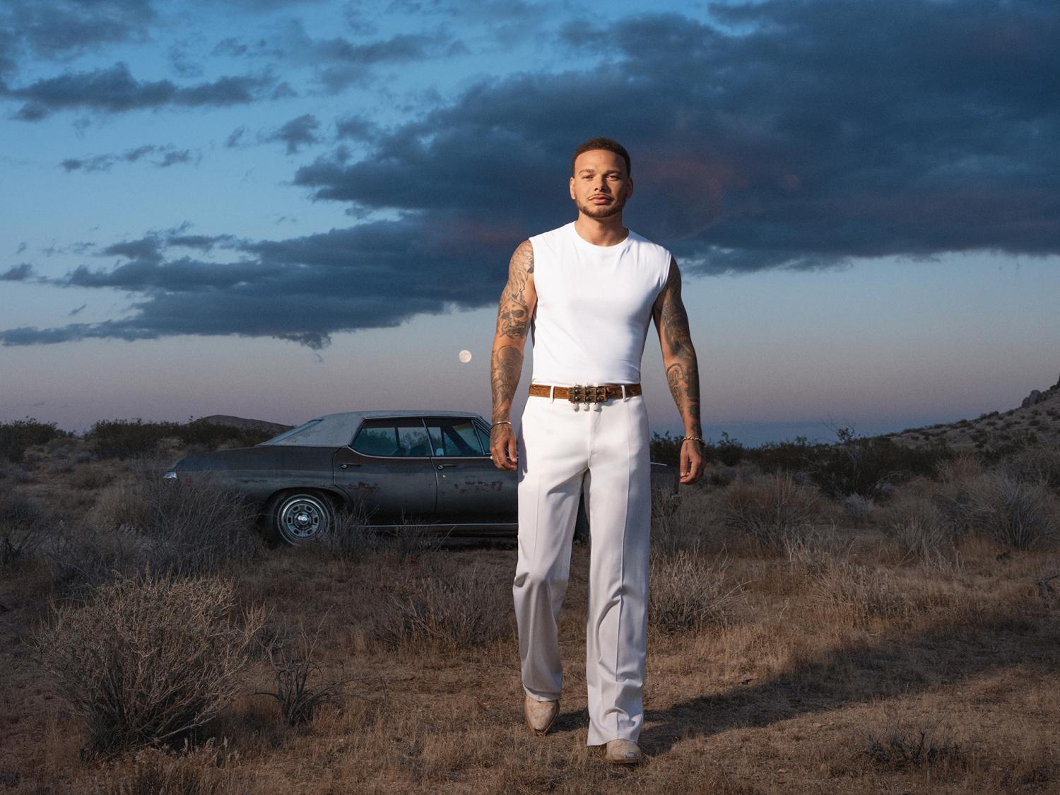 Kane Brown in front of a car in the country.