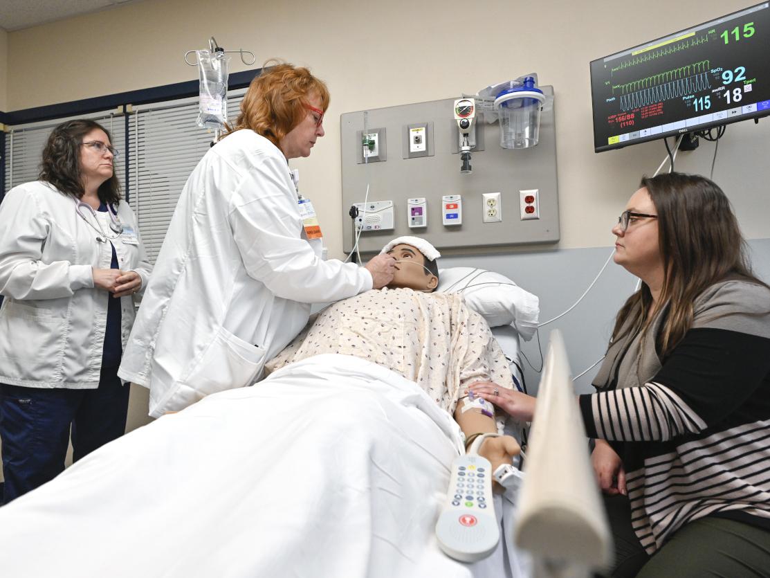Two nurses tend to a mannequin posing as a patient while a woman sits at the bedside, looking on.