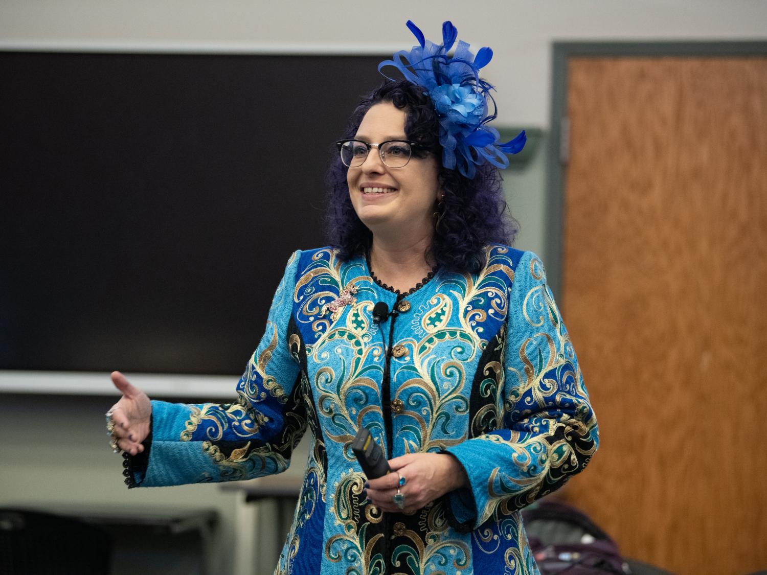 A person wearing a blue fascinator stands in front of a blackboard and a door.
