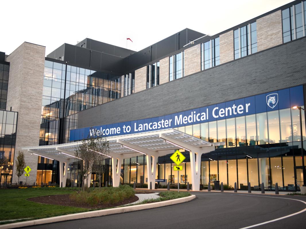 Front entrance of Penn State Health Lancaster Medical Center. The building features a sleek design with extensive glass windows, beige and dark gray brickwork, and a covered entrance with white, curved support beams. A pedestrian crossing sign is visible near the driveway, surrounded by green landscaping and trees.