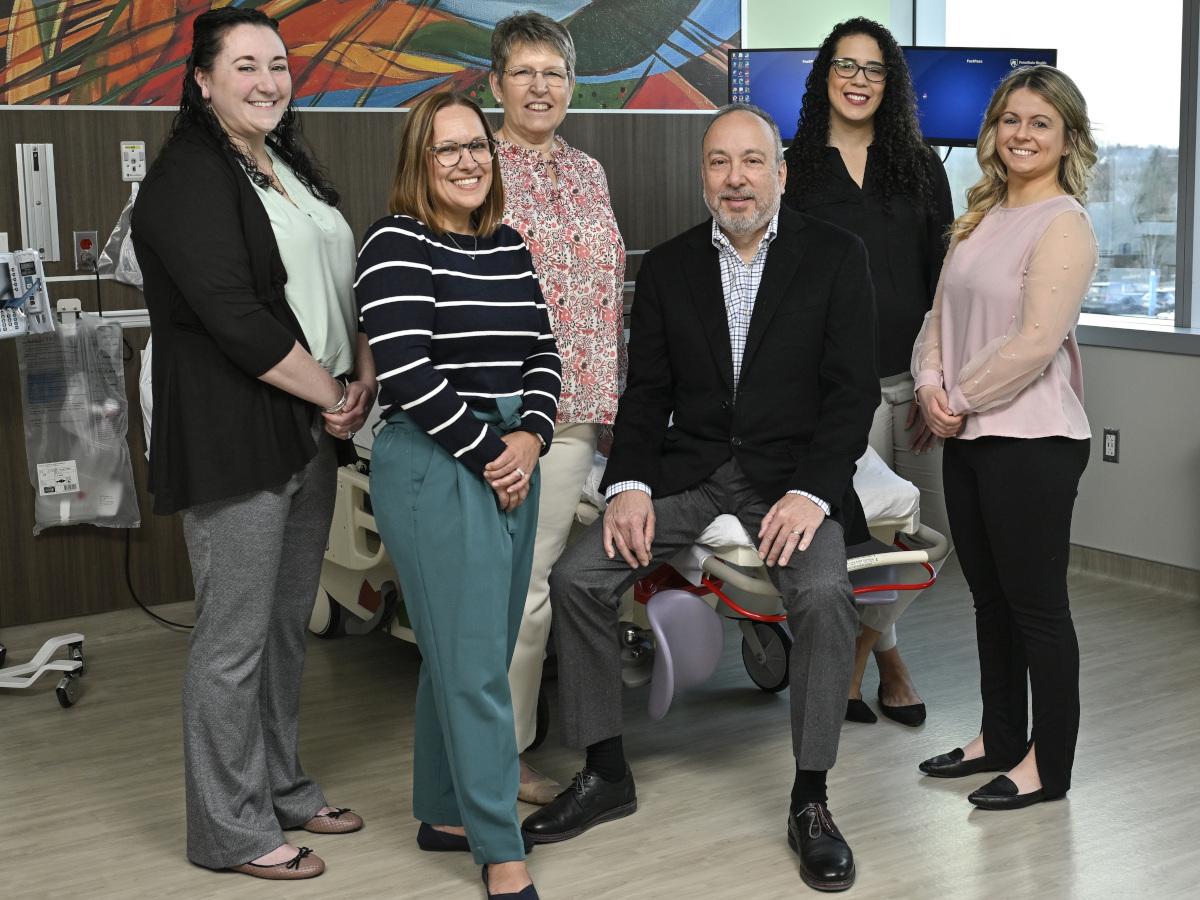 Six people in business casual attire pose for a photo in a hospital room. A hospital bed, computer and other medical equipment are in the background.
