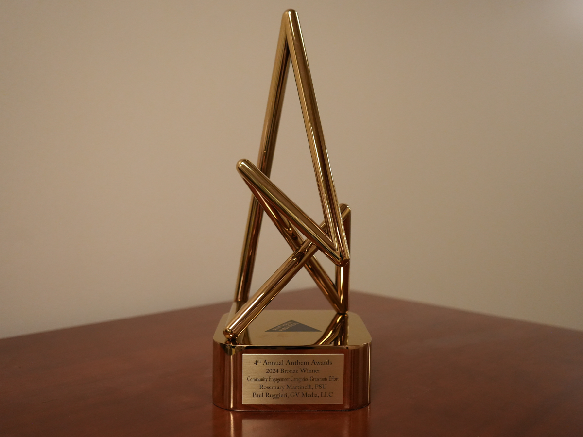 A bronze anthem award on a table, the words "4th Annual Anthem Awards 2024 Bronze Winner; Community Engagement Categories-Grassroots Effort; Rosemary Martinelli, PSU; Paul Ruggieri, GV Media, LLC" are engraved upon its base.