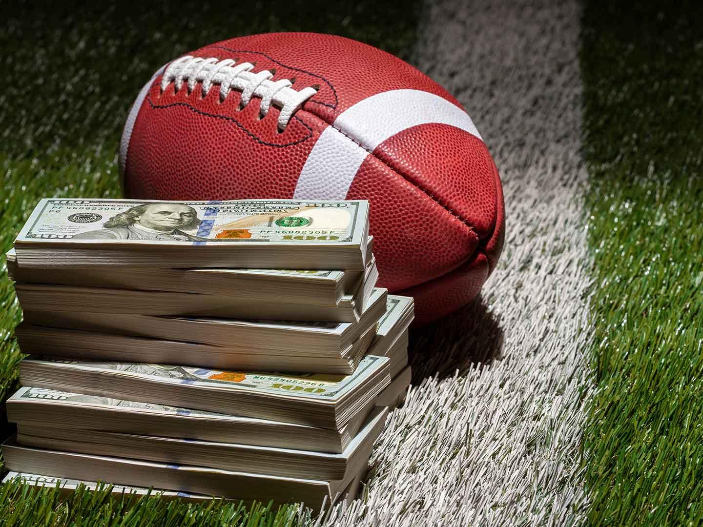 A football and stacks of one hundred dollar bills sitting on green turf grass