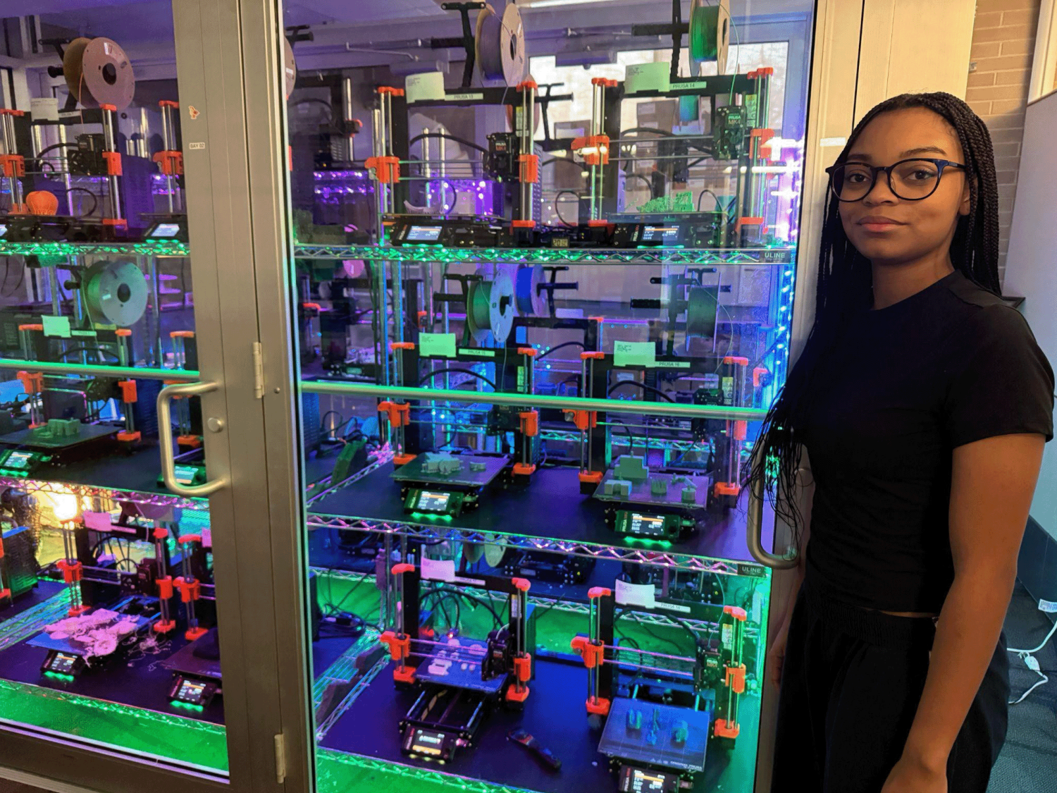 Penn State student and Maker-in-Residence, Keyana Gales-Cook, stands next to a large, glass-enclosed rack filled with multiple 3D printers. She is wearing glasses and a black T-shirt, looking confidently into the camera. The brightly lit 3D printers are actively printing objects, showcasing the university’s 3D printing service.