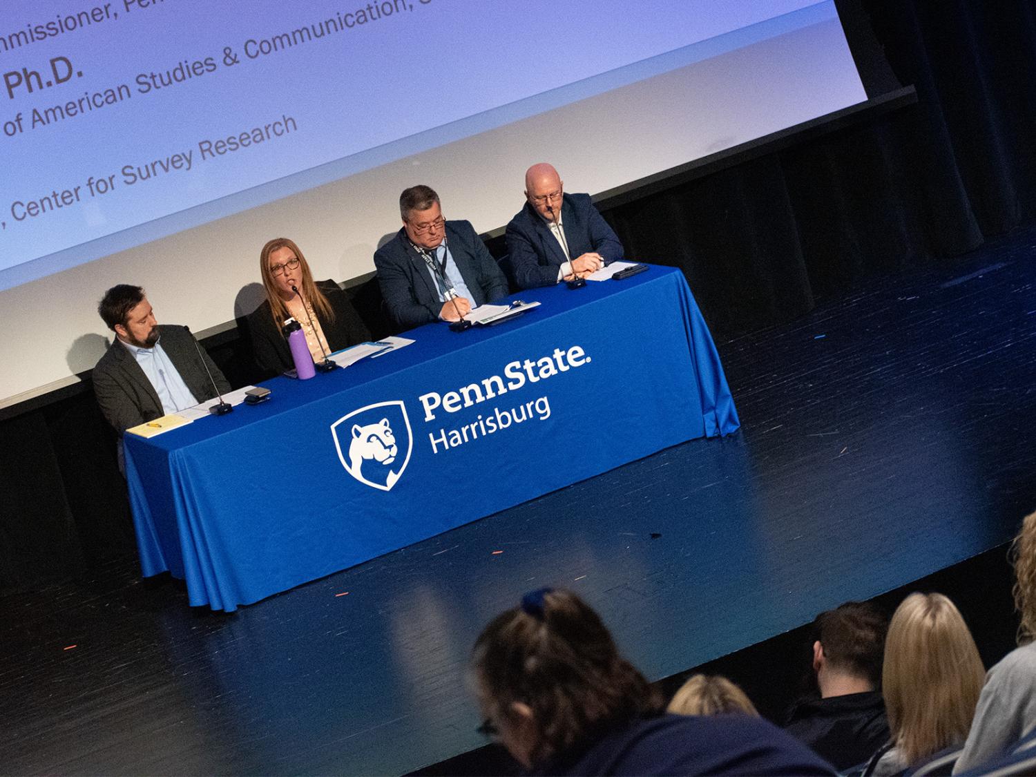 A photo of four people sitting behind a table during a presentation on a stage