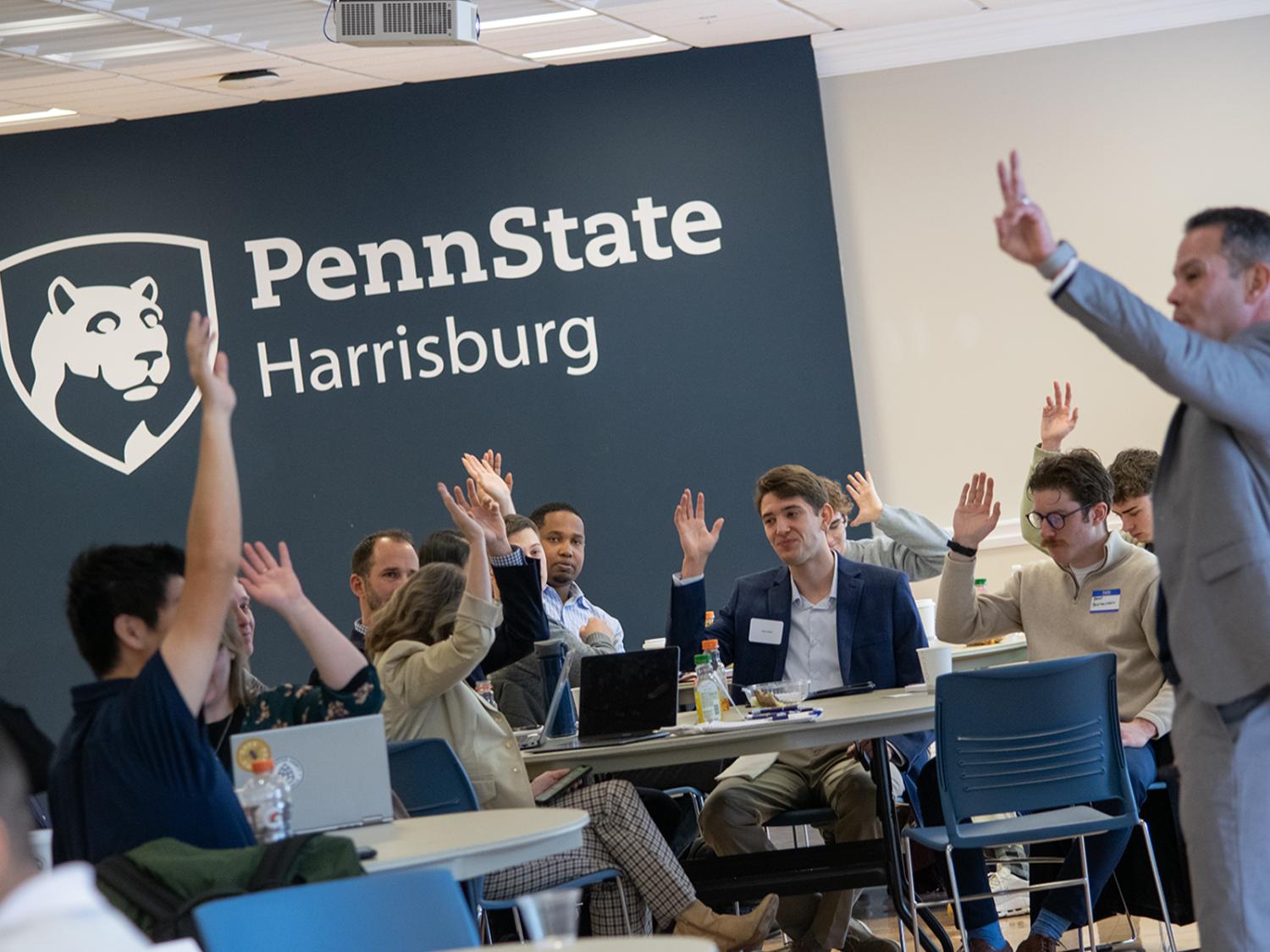 People sitting at tables raise their hands, the Penn State Harrisburg logo on the wall behind them