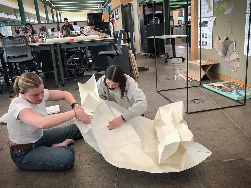 Two students sit on the ground in a lab working on folding a stiff paper origami mold.