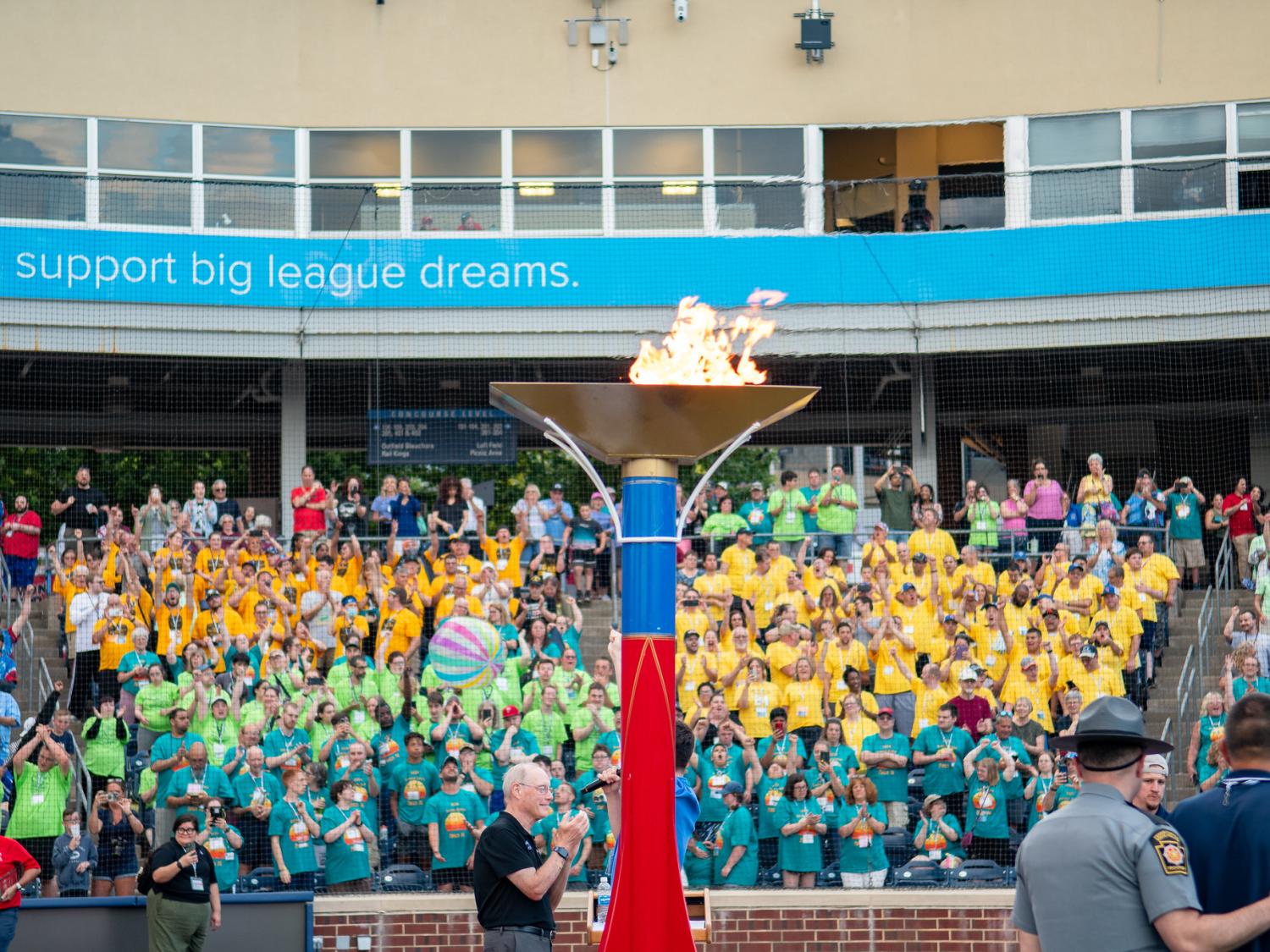 A huge lit torch is backgrounded by stadium stands full of people wearing colorful t-shirts