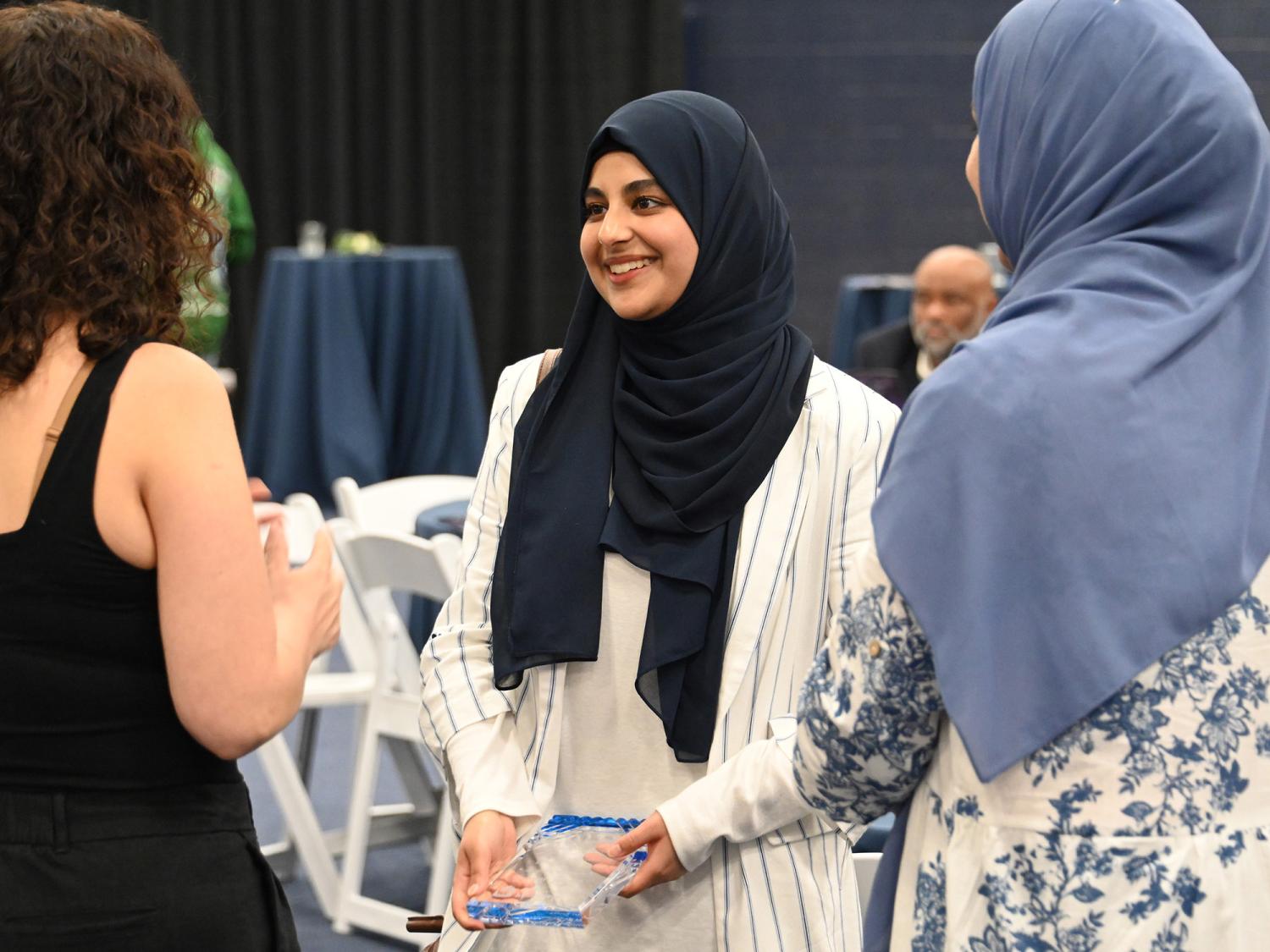 Three students talk and smile during the Evening of Excellence celebration at Penn State Greater Allegheny. One holds a clear award plaque.