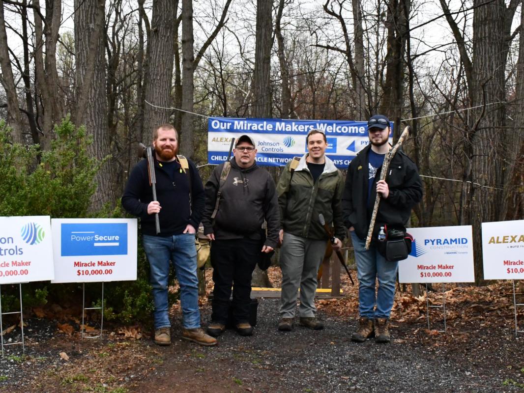 Four men pose for a photo at the edge of a wooded area. Three of them hold guns pointed at the ground.
