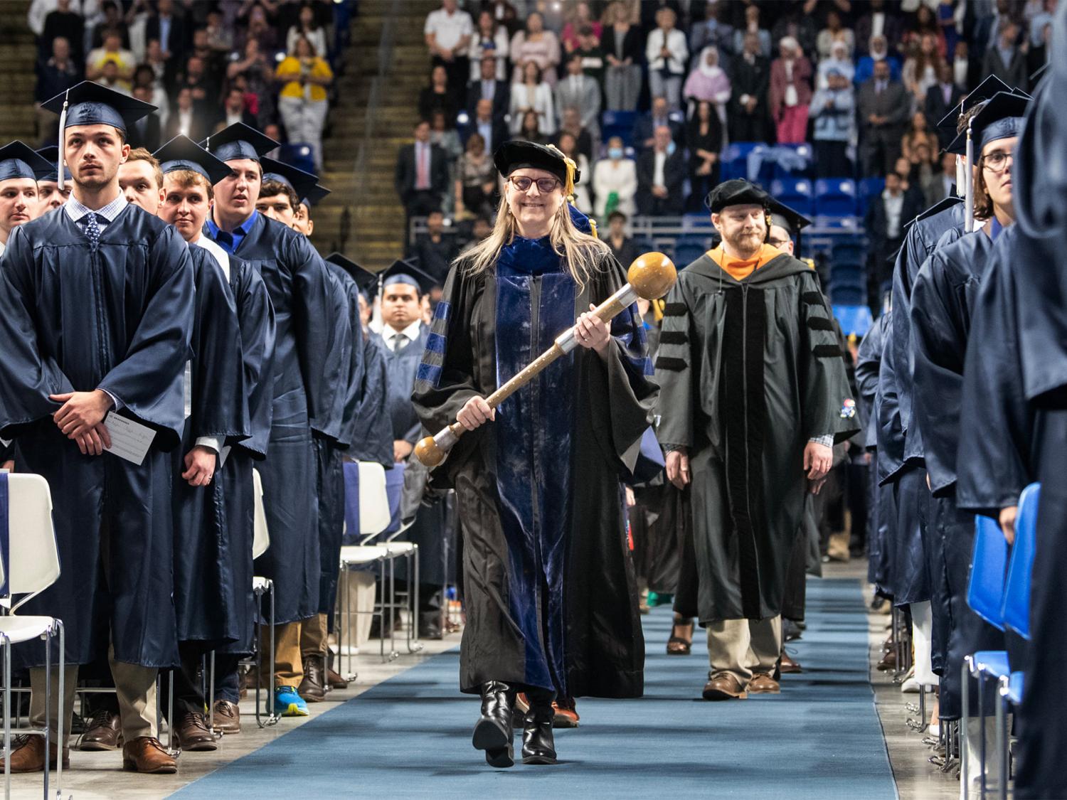 Person walking down aisle at commencement, surrounded by others watching on.