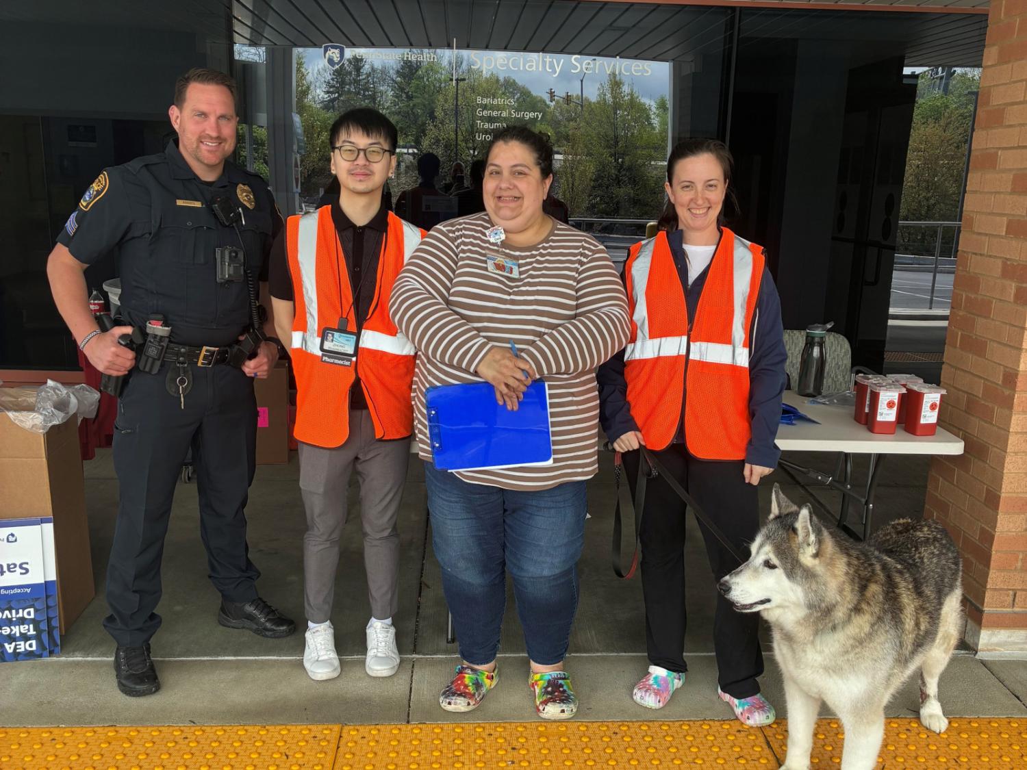 A police officer, three colleagues and a dog stand next to tables and collection boxes in front of a medical practice.