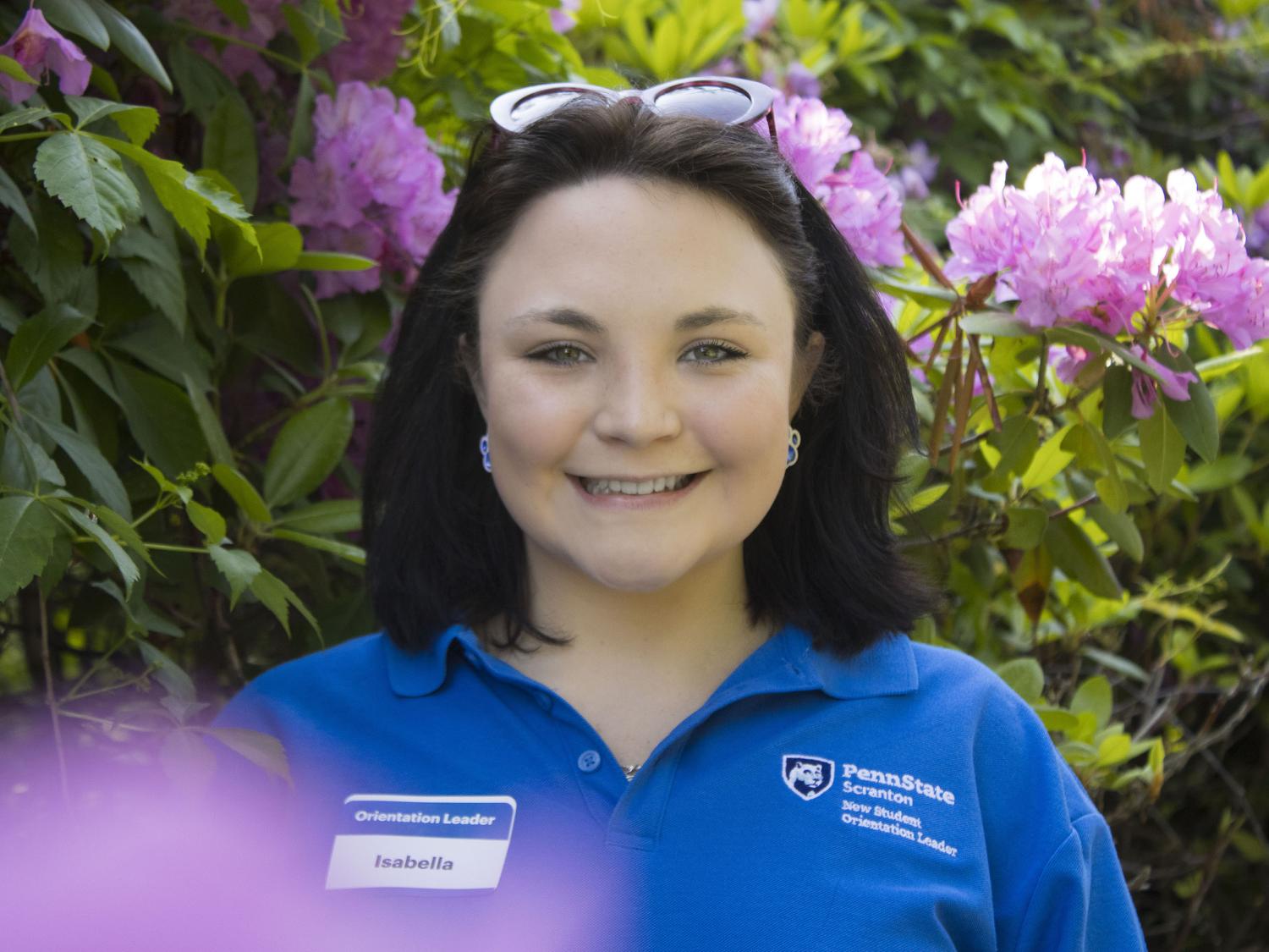Student smiles in front of flowers wearing a shirt that reads 'New Student Orientation'