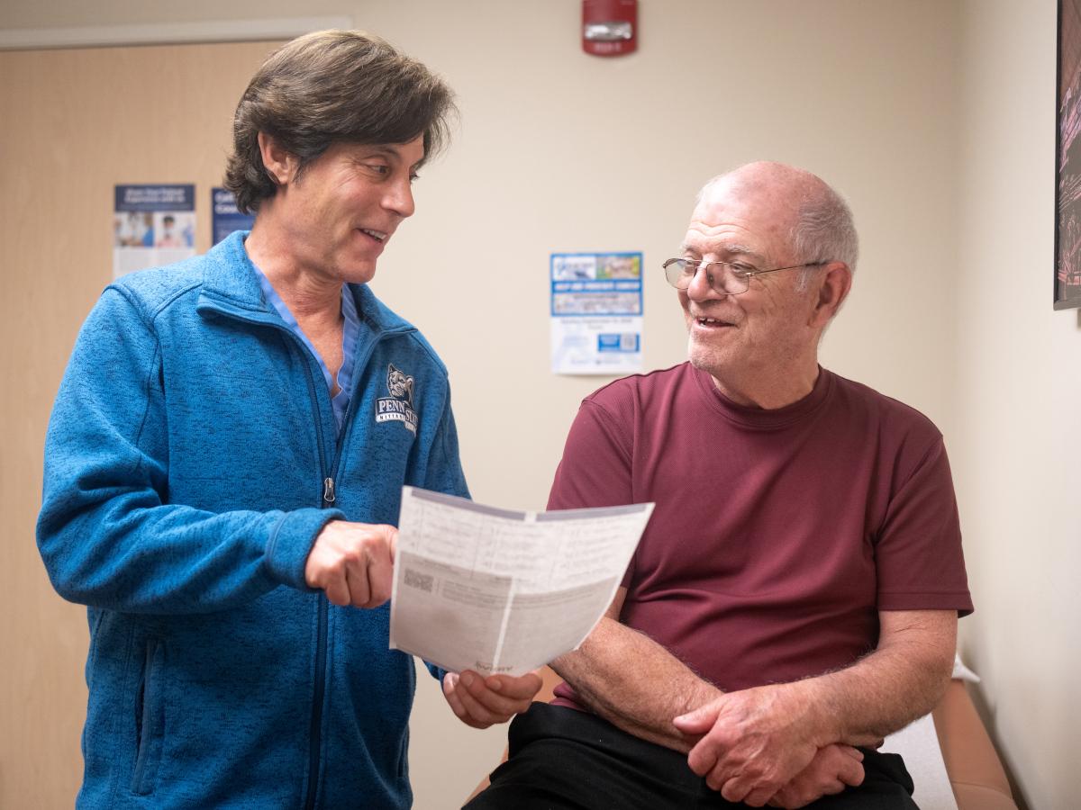 A doctor smiles at a male patient as he points to a paper he is holding. The doctor is wearing a sweat jacket with a Penn State logo on it. The male is wearing a T-shirt and glasses.