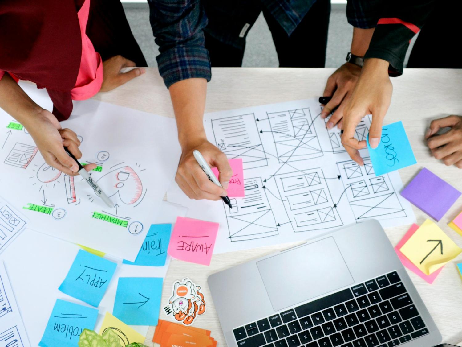 Aerial view of people's hands working on project with diagrams, sticky notes, and a laptop.