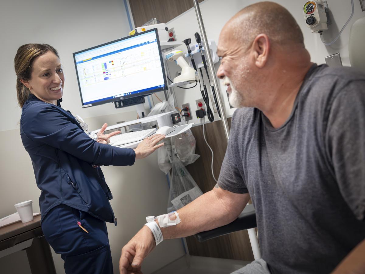 A nurse stands at a computer, looking to the side as she speaks with a patient. They are in a hospital emergency room treatment bay.