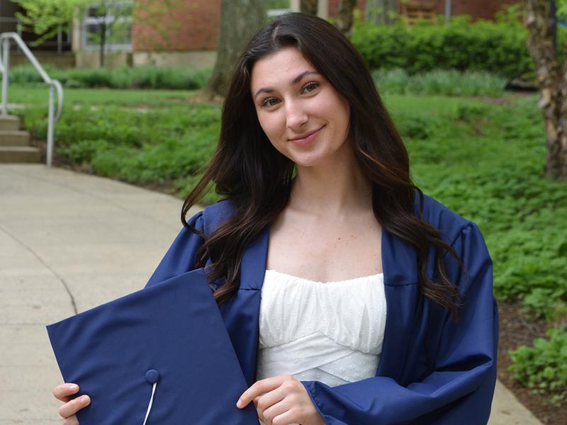 Young woman poses in front of Chambers Building wearing graduation gown and holding cap to her right.