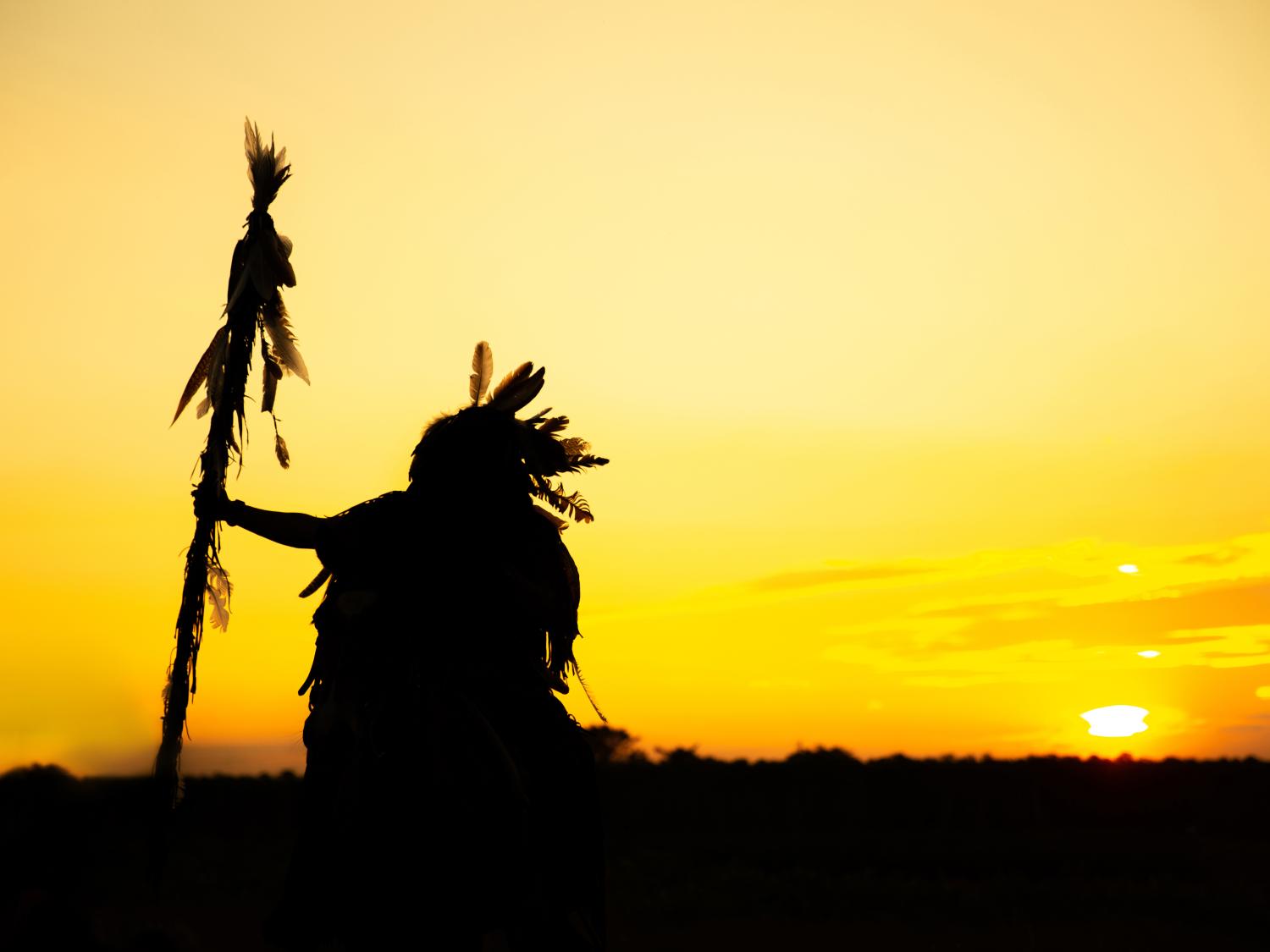 sunset silhouette of person wearing feathers on head and holding tall feathered staff