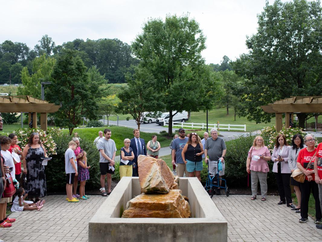 Approximately 20 people stand on a brick patio, surrounding a stone memorial, at a ceremony.