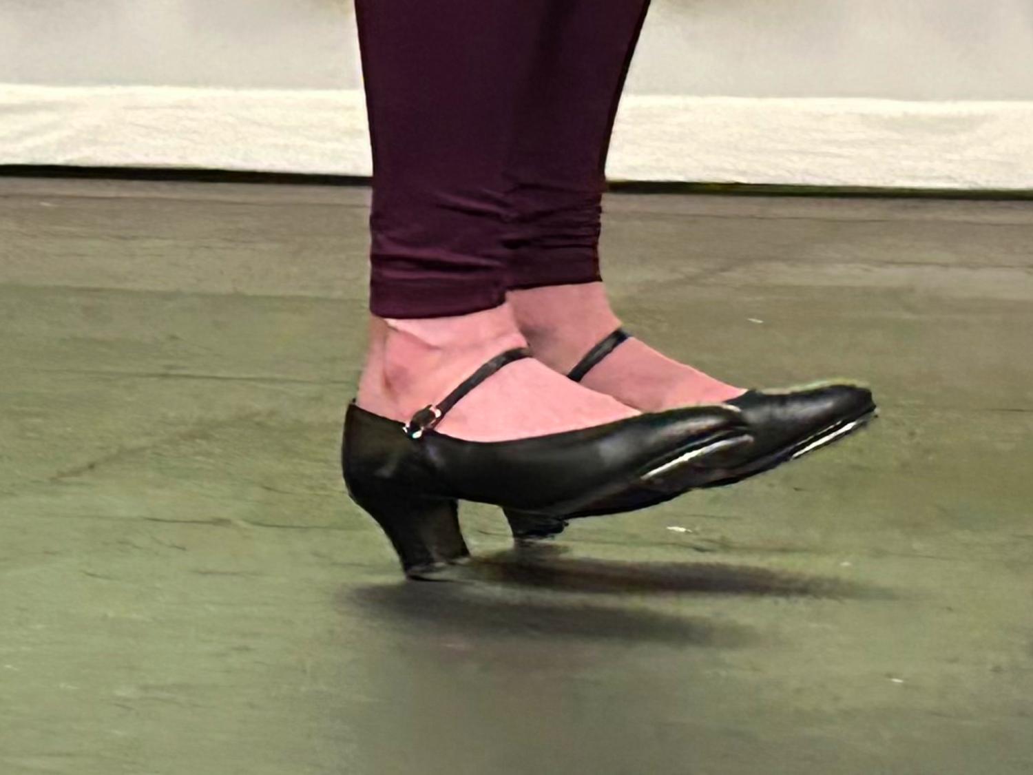 A close-up photo of a dancer's feet mid-air, wearing black character tap shoes with a low heel and ankle straps.