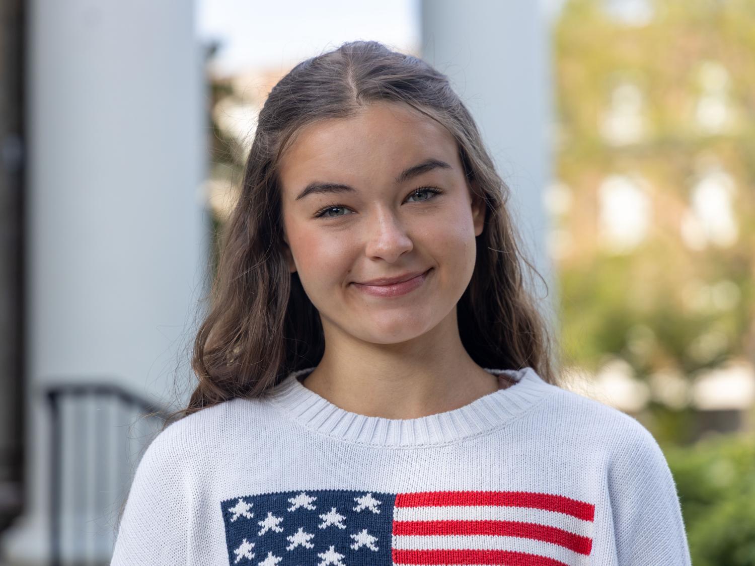 Isabelle Ems wears a white sweater with an American flag on it in front of a backdrop of columns.