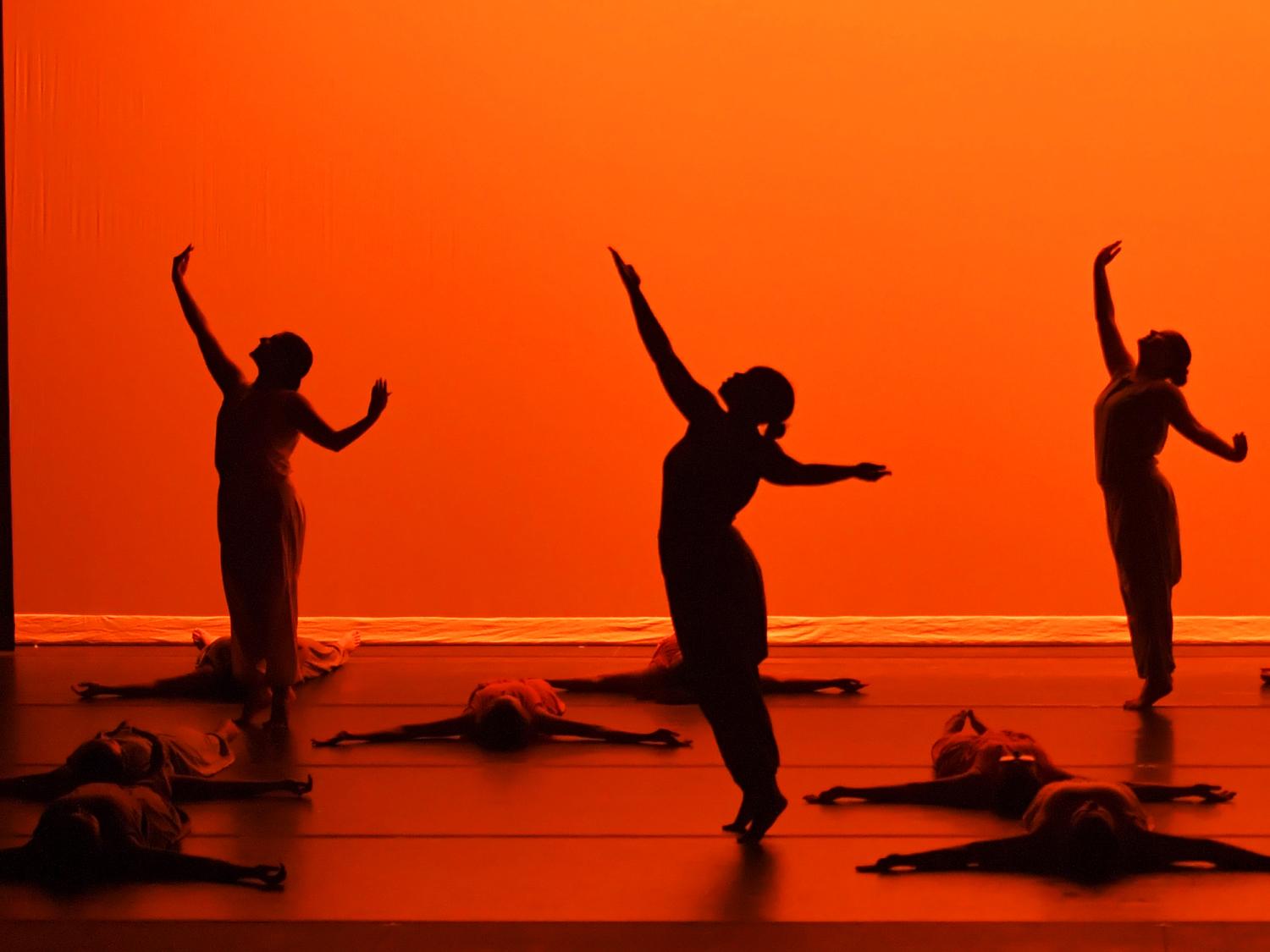 Silhouetted dancers pose gracefully on stage against a glowing orange backdrop, with others lying on the floor.