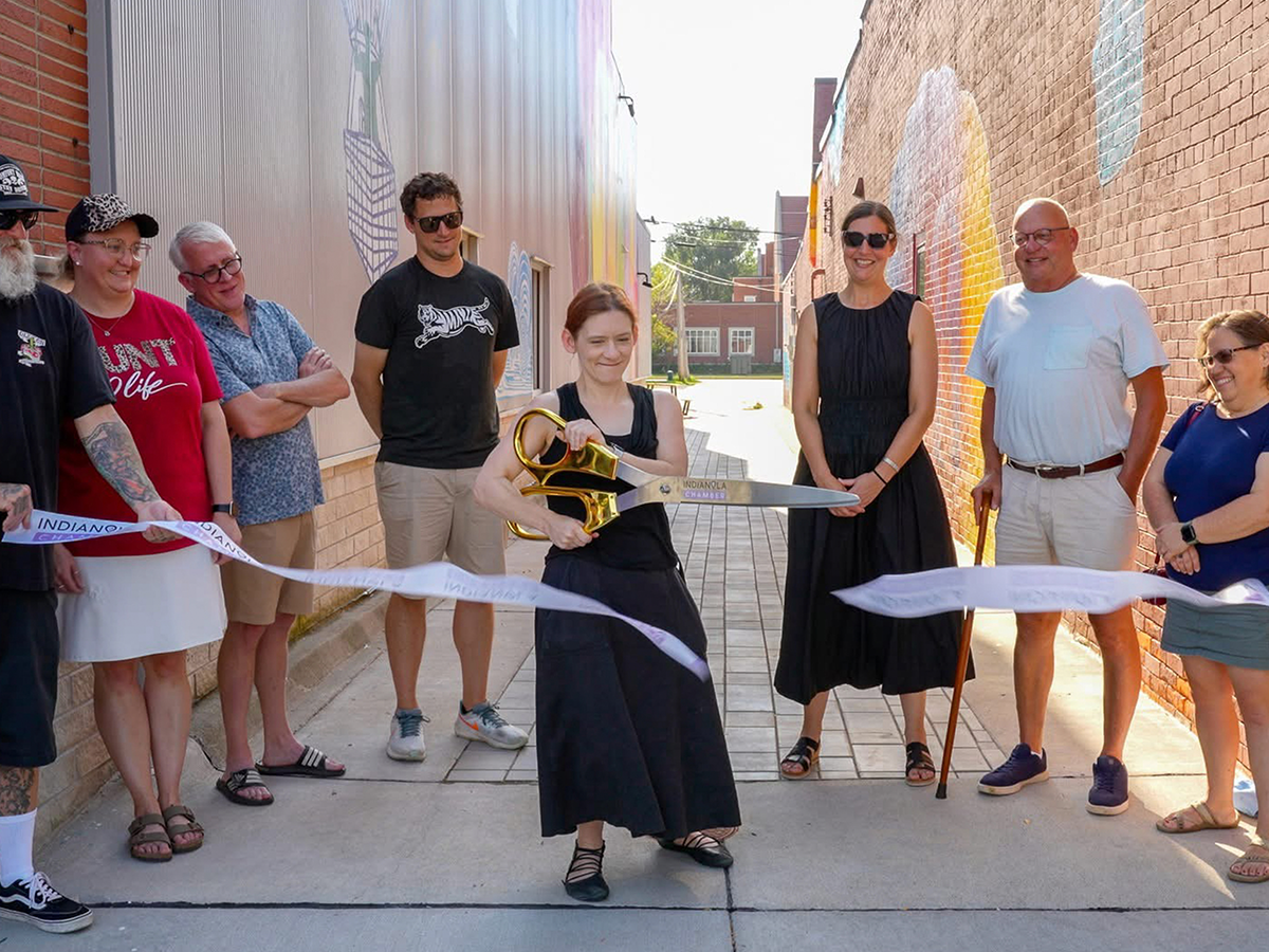 Lori Hepner cuts a ceremonial ribbon with oversized scissors at the entrance of Buxton Alley in Indianola, Iowa, while community members stand smiling on either side of her.
