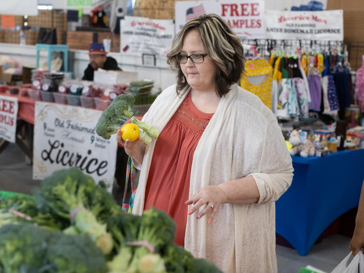 A woman holds a head of broccoli and an orange while looking at other produce at a farmer’s market. She is wearing a sweater and has glasses. 