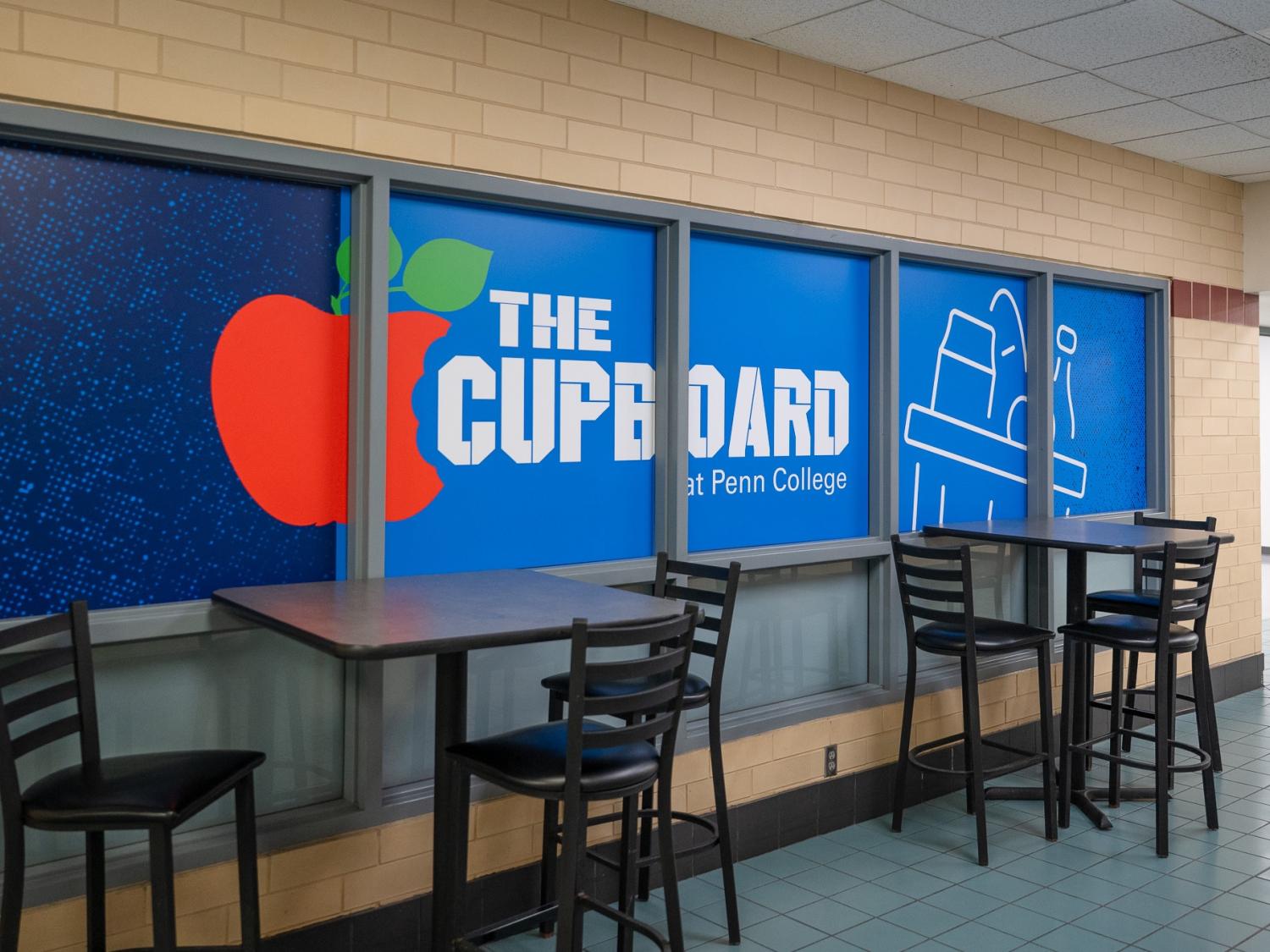 Tables in chairs next to a blue wall that says "The Cupboard at Penn College" with an apple illustration.