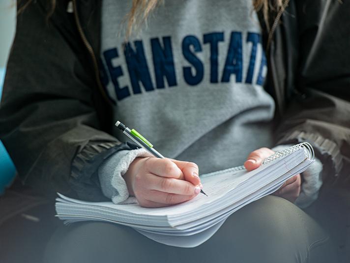A student wearing a Penn State shirt takes notes in a notebook.