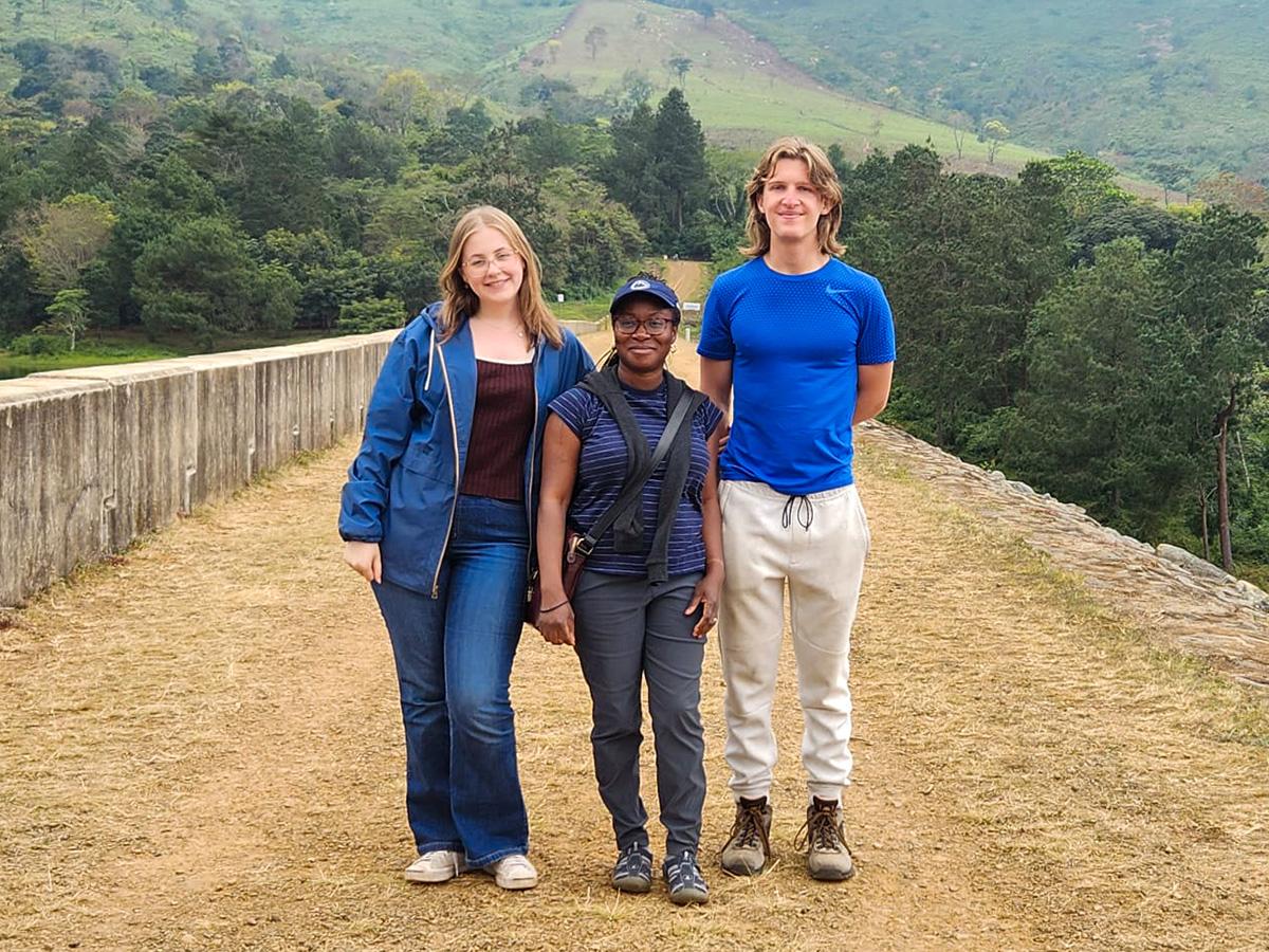 Three people stand on a dirt pathway with lush green hills in the background in Malawi.