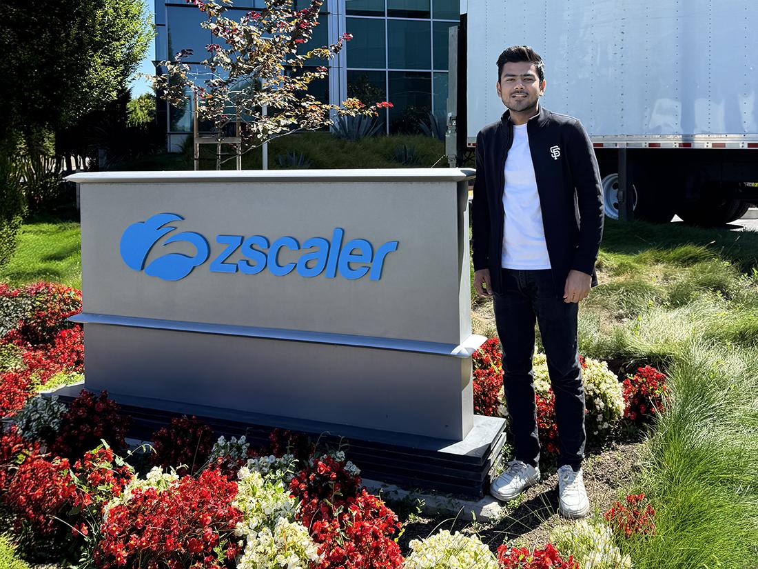 An intern stands outside a corporate sign for the company Zscaler